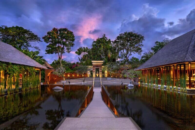 A stone pathway crosses a reflective pond, leading to a modern building at Amarterra Villas Nusa Dua, surrounded by lush trees and tropical architecture at dusk.