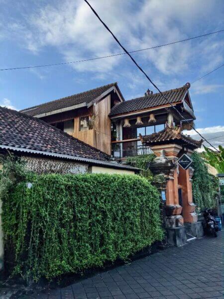 Sunshine Vintage House is a two-story traditional Balinese building in Ubud with a tiled roof, brick gate, hanging plants, and a dense green hedge along a paved street under a blue sky.