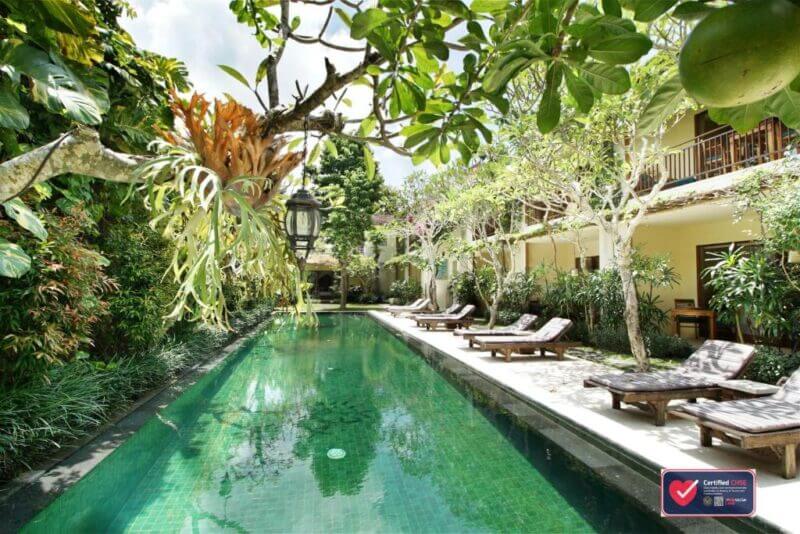 Outdoor swimming pool lined with lounge chairs and lush greenery at Sapodilla Ubud, adjacent to a two-story building with balconies.