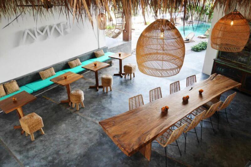 Modern open-air cafe at Amar Boutique Hotel with natural wood tables, wicker chairs, and hanging woven lights. A turquoise bench lines one wall, and a pool is visible outside—perfect for unwinding in the heart of Canggu.