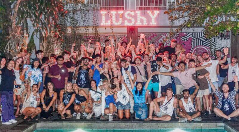 A large group of people pose together in front of a building with a neon LUSHY sign at Lushy Hostel Canggu, some gesturing energetically, beside a pool at night.