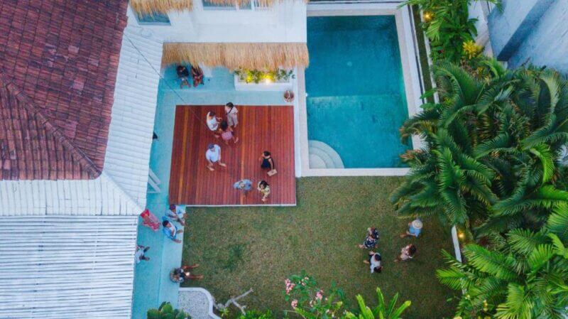Aerial view of people gathering on a wooden deck and lawn near a swimming pool, surrounded by tropical plants and white buildings.