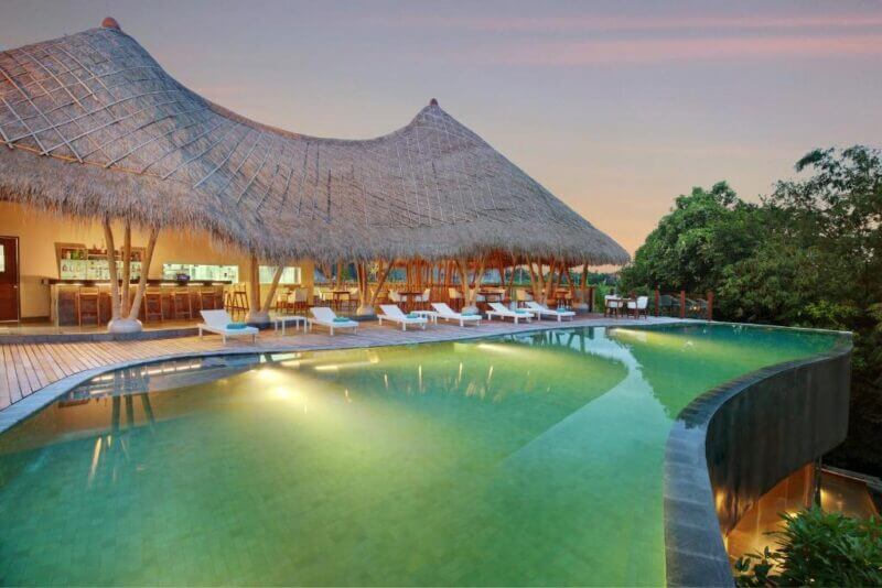 Infinity pool with sun loungers and a thatched-roof open-air bar at sunset, surrounded by trees, at Theanna Villa and Spa Canggu.