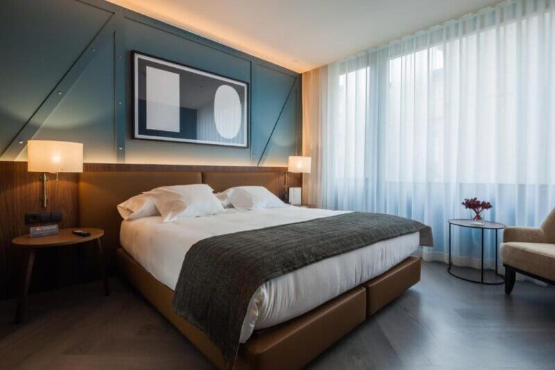 Modern Porto Vincci hotel room with a double bed, two bedside lamps, abstract wall art, floor-to-ceiling window with sheer curtains, round side table with flowers, and an inviting armchair.