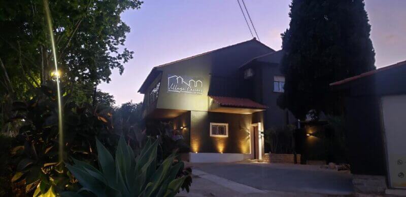 A modern building with a lit Village Casual sign, surrounded by greenery in Village Cascais, at dusk with outdoor lights illuminating the entrance.