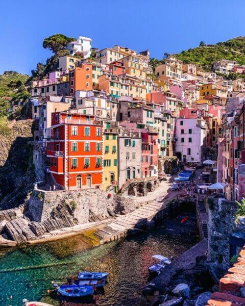 Colorful buildings of Cinque Terre line a cliffside above the small harbor with boats in clear water under a blue sky, near the charming area of LA RIPA in Riomaggiore, Italy.