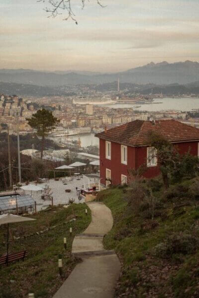 A winding path leads past a red house on a hillside in Cinque Terre, overlooking a city and harbor, with mountains visible in the background under a cloudy sky.