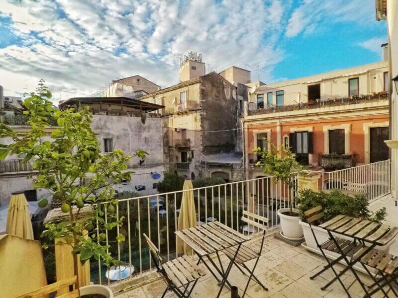The outdoor balcony at La Residenza Siracusa, with potted plants and empty metal chairs and tables, overlooks old buildings under a partly cloudy blue sky—an inviting scene for your Sicily accommodation.