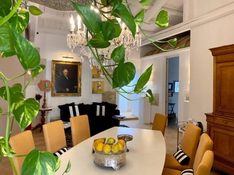 A dining room at La Maison Siracusa features a white oval table, yellow chairs, a crystal chandelier, framed portraits on the wall, and a leafy plant in the foreground. A bowl of fruit sits on the table.