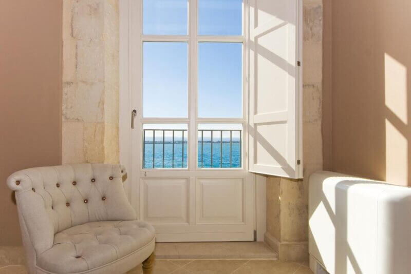 A white cushioned chair sits near a tall window with open shutters at Palazzo Alfeo in Siracusa, overlooking a clear blue sea under a sunny sky.