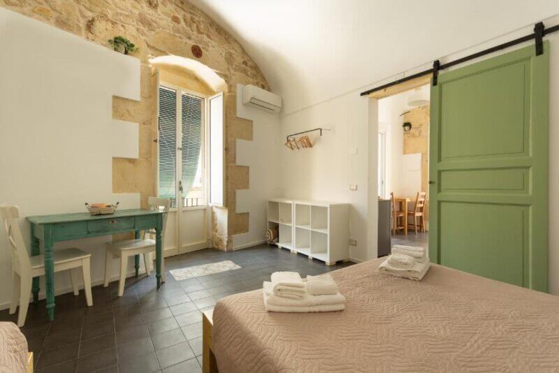 Bright bedroom with stone accent wall, a green sliding door, twin beds with folded towels, a small desk, and an open window letting in natural light—perfect for enjoying a Pura Vida vibe in Ragusa.
