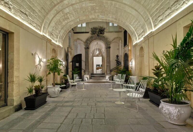 Charming stone courtyard at 1768 Boutique in Ragusa, featuring an arched ceiling, lush potted plants, and white metal chairs and tables leading to a welcoming building entrance.