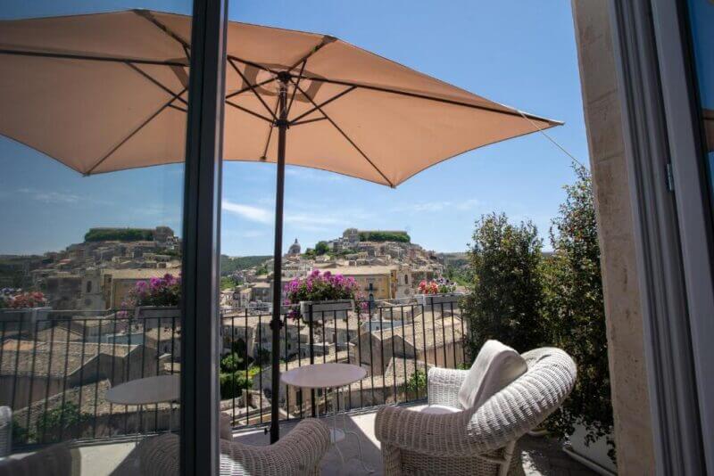 Outdoor balcony with two white wicker chairs, a small round table, potted flowers, and a large umbrella, overlooking the picturesque hilly townscape of Ragusa under a clear blue sky.