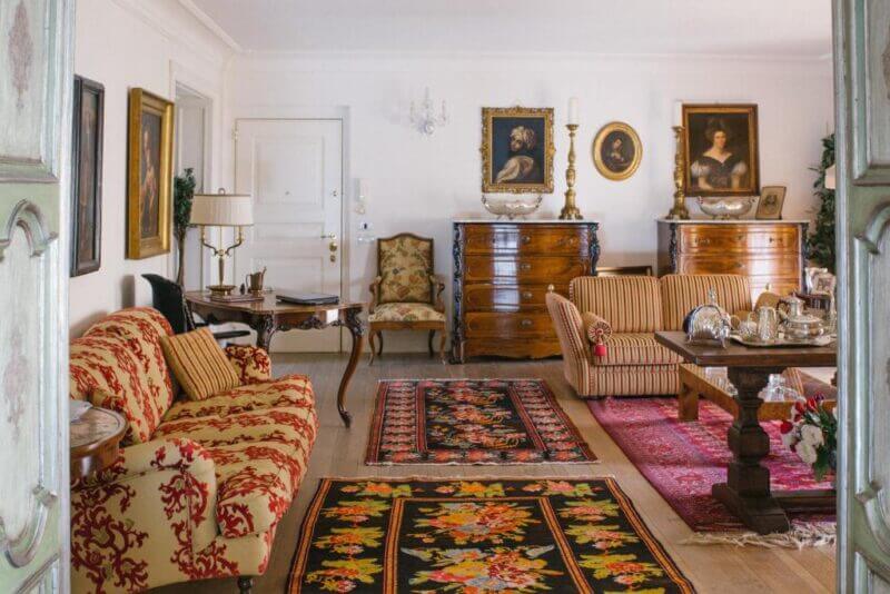 A traditional living room with ornate patterned sofas, wooden furniture, framed portraits, floral rugs, and decorative items—some inspired by Agrigento’s heritage—adorn tables and chests beneath a subtle Triskéles motif.
