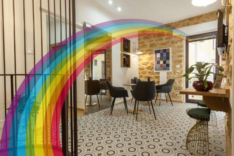 A modern dining room at Casa Tita features patterned tile floors, stone walls, black chairs, a round table, and a large rainbow graphic superimposed across the image—bringing a touch of Agrigento’s charm indoors.