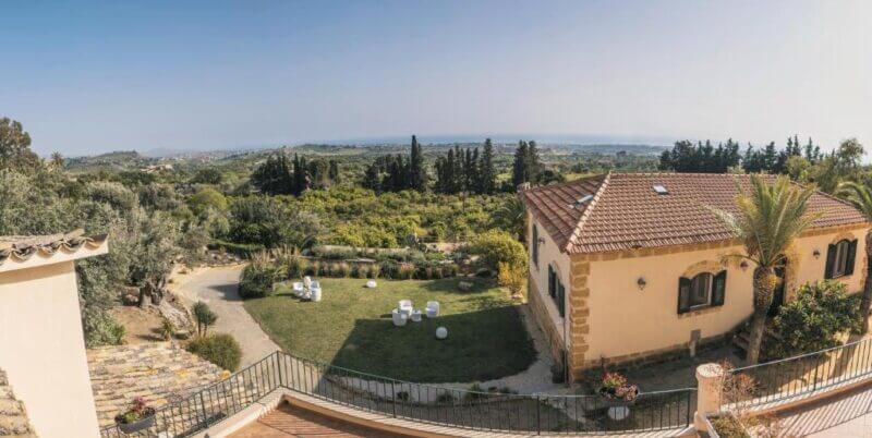 A panoramic view of Villa Lumia in Agrigento, featuring a tiled-roof house, a well-kept yard with outdoor seating, lush greenery, trees, and a distant landscape beneath a clear sky.