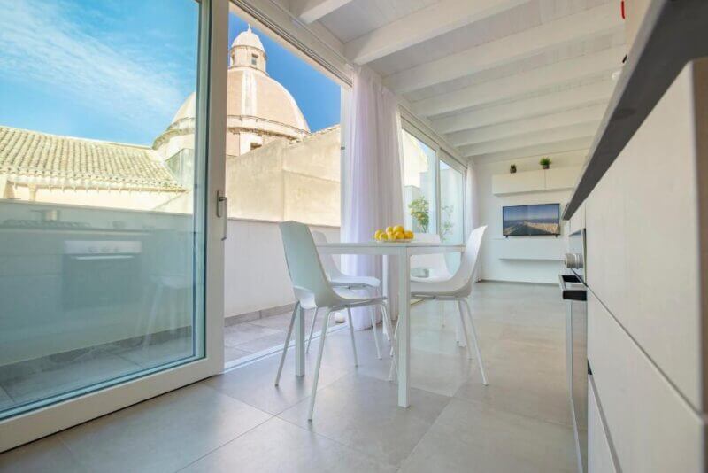 Modern kitchen and dining area with white furniture, large windows, and a view of Le Cupole Trapani’s iconic domed building outside.