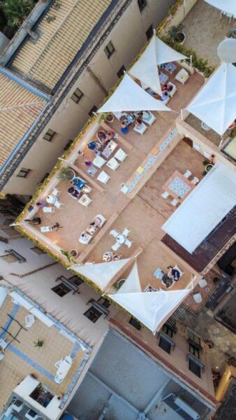 Aerial view of BADIA NUOVA’s rooftop terrace in Trapani, with people sitting at tables, surrounded by buildings and white shade sails overhead.