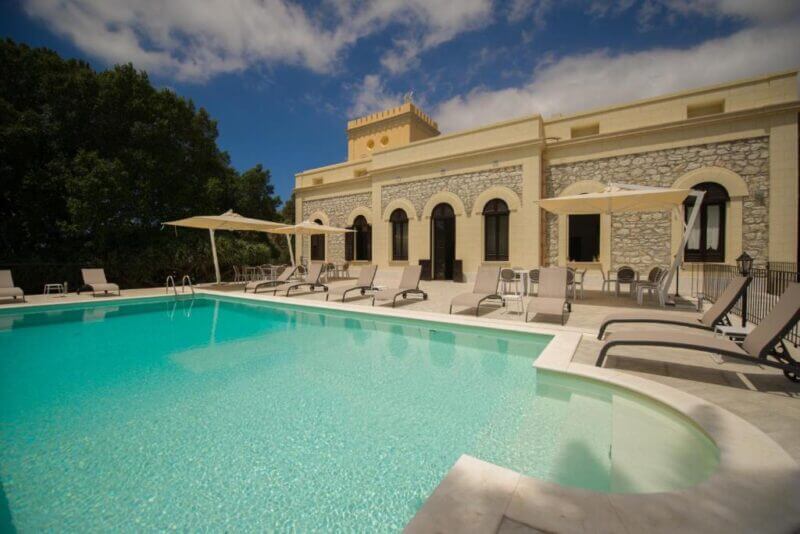 Outdoor swimming pool with lounge chairs and umbrellas in front of Villa le Torri Trapani's beige stone building under a partly cloudy sky.