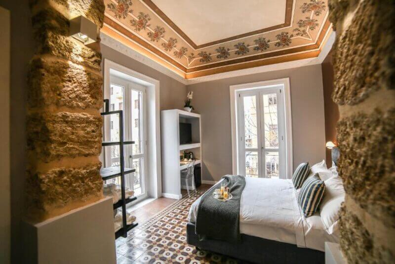 A modern Family Affair Palermo bedroom with patterned floor tiles, a decorated ceiling, a double bed, a desk with a TV, and large windows letting in natural light.