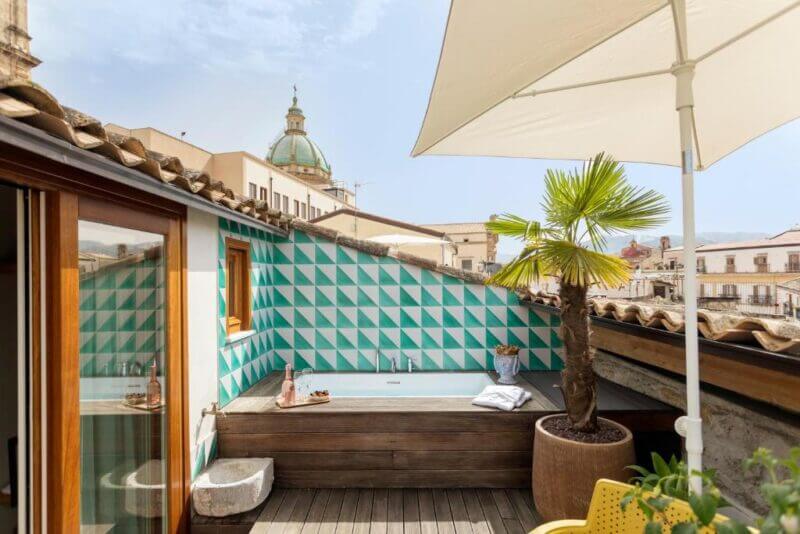 Rooftop terrace at Sui Tetti Palermo with a jacuzzi, geometric tiled wall, potted palm, umbrella, wooden deck, and city dome in the background.