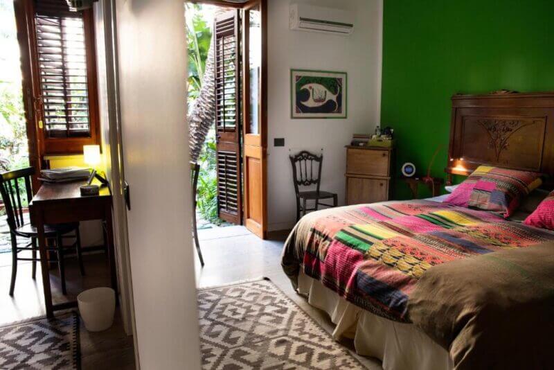 A cozy bedroom with a colorful bedspread, green accent wall, wooden furniture, and a desk captures the charm of Cefalu. Open doors lead to a garden as natural light fills the Lirma-inspired retreat.