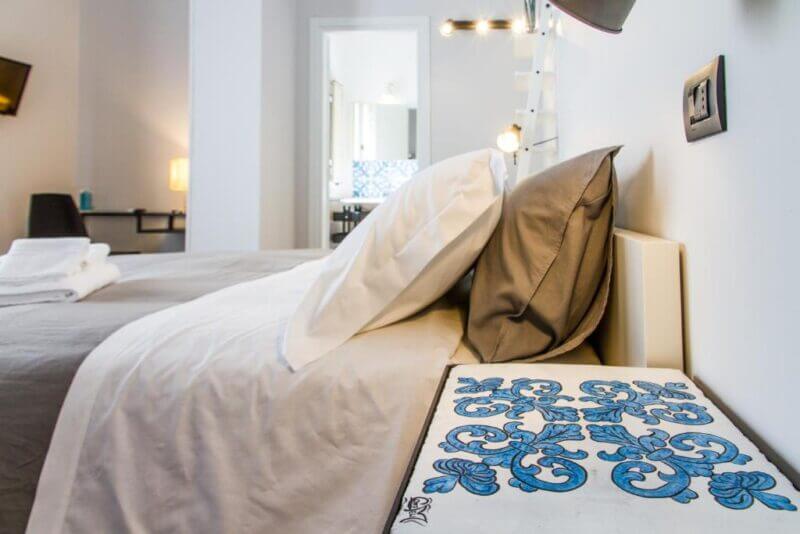 Modern bedroom with neatly made bed, gray and white bedding, white walls, a decorative blue bedside tile, and a view into a bright bathroom—perfect for your stay at Cortile Umberto in charming Cefalu.