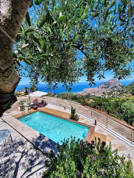 A rectangular swimming pool with a wooden deck at Al 33 Giri Cefalu overlooks a coastal town and the sea, surrounded by hills and greenery, all viewed through tree branches.