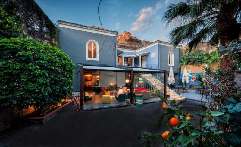 A two-story blue house with large windows, an outdoor seating area, and stairs leading to a balcony, surrounded by lush greenery and citrus trees—experience comfort and relaxation at Yard Hostel Catania.