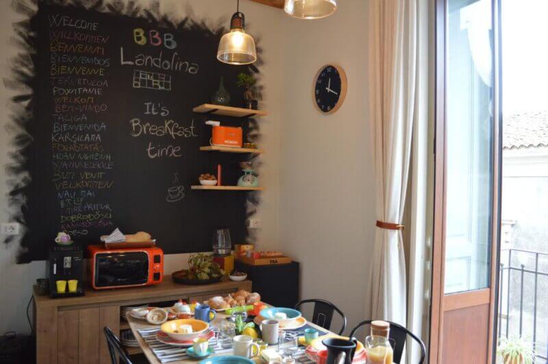 A cozy breakfast room at Landolina in Catania features a set table, a chalkboard wall welcoming guests in multiple languages, charming decor on shelves, and a door leading to a sunny balcony.