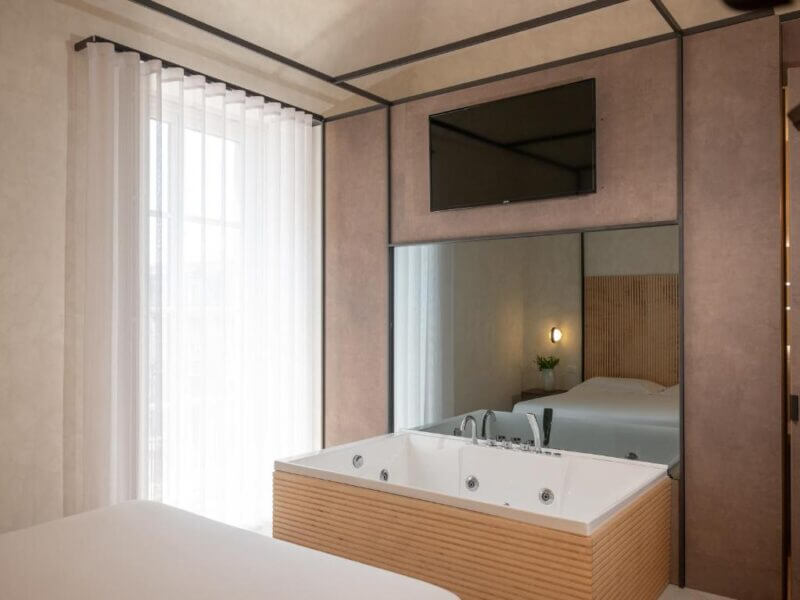 Modern hotel room at Dome Catania featuring a built-in jacuzzi tub, a wall-mounted TV above a large mirror, sheer curtains over the window, and part of a bed in view.
