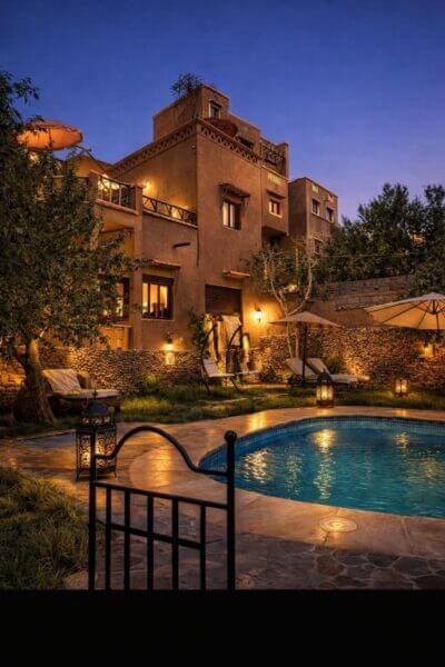 A multi-story Riad with warm lighting overlooks a round swimming pool surrounded by lounge chairs, trees, and lanterns at dusk, evoking the enchanting ambiance of the Sahara.