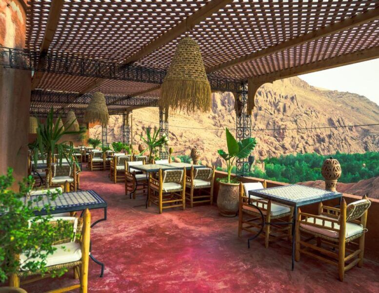 Outdoor restaurant terrace at Riad Rihana Dades with wooden tables and chairs, woven pendant lights, potted plants, and a view of rocky mountains and greenery in the background.