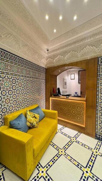 A yellow sofa with blue and yellow pillows sits in front of a reception desk, surrounded by ornate patterned tiles and detailed plasterwork, capturing the charm of traditional Chefchaouen accommodation.