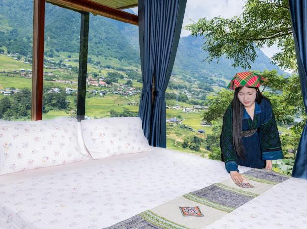 A woman in traditional clothing arranges bedding on a large bed near a window inside a Hmong house, with views of green hills, trees, and the village of Sapa outside.