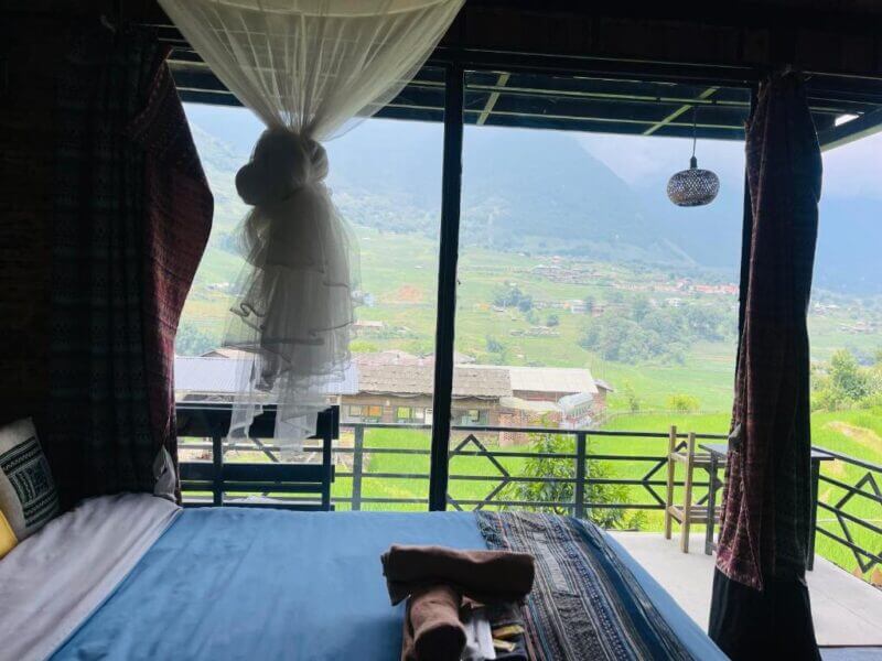 A bed with folded towels and a canopy faces open curtains and a balcony at Plum Eco House Sapa, overlooking green fields and mountains under a cloudy sky—an inviting, eco-friendly accommodation in the heart of Sapa.