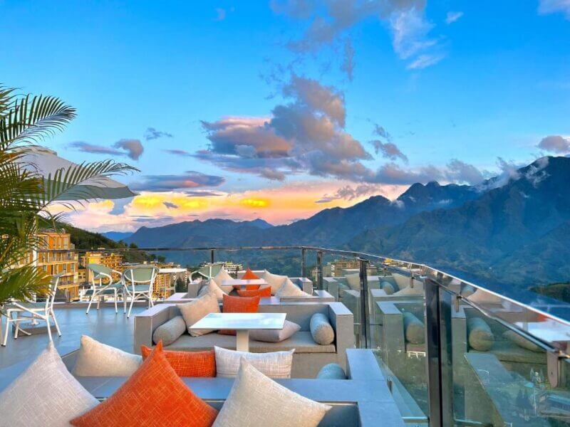 Outdoor rooftop lounge at Relax Hotel Sapa with cushioned seating and tables, overlooking a stunning Sapa mountain landscape at sunset under a partly cloudy sky.