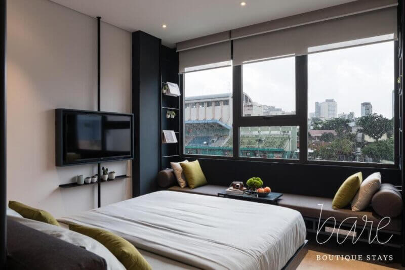Modern bedroom with a large bed, wall-mounted TV, and a window overlooking Ho Chi Minh’s cityscape and sports field; Bare Chic boutique stays logo in the corner.