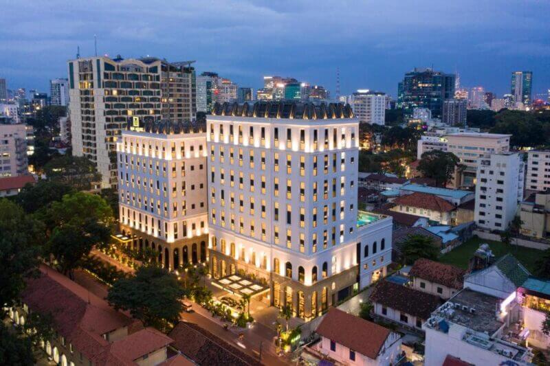 A tall, modern hotel building—Mai House Ho Chi Minh—with illuminated windows stands among trees and surrounding city structures at dusk. High-rise buildings and vibrant city lights are visible in the background.