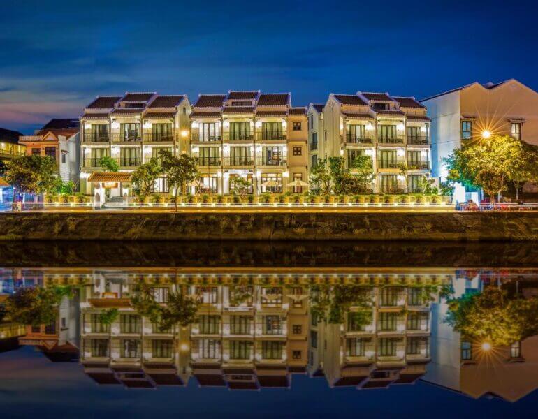 Row of illuminated buildings at night, including a charming riverside hotel, with their reflections shimmering in the calm water of Hoi An’s canal.