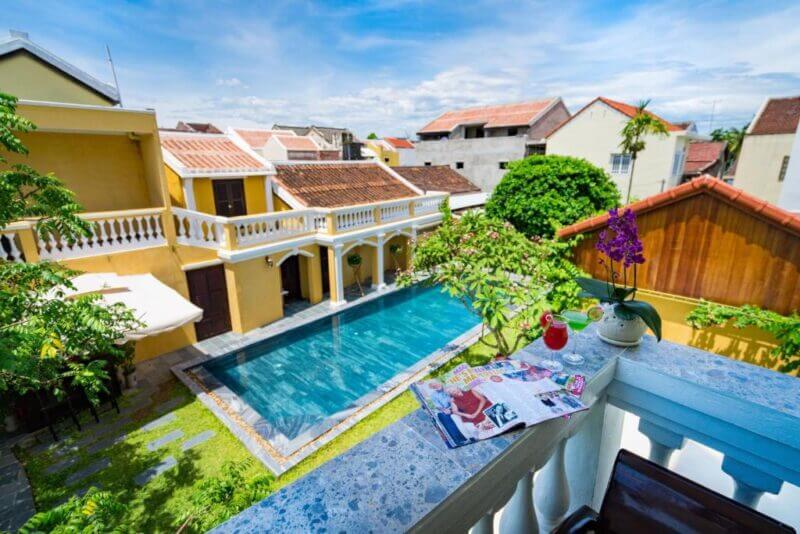 View of a rectangular outdoor swimming pool at a central hotel in Hoi An, surrounded by yellow buildings, seen from a balcony with magazines, a pink drink, and a potted orchid on the railing.