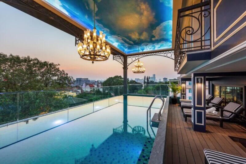 Rooftop swimming pool at Aira Boutique Hanoi hotel with chandeliers, painted ceiling, lounge chairs, wooden deck, and a stunning cityscape view at sunset.