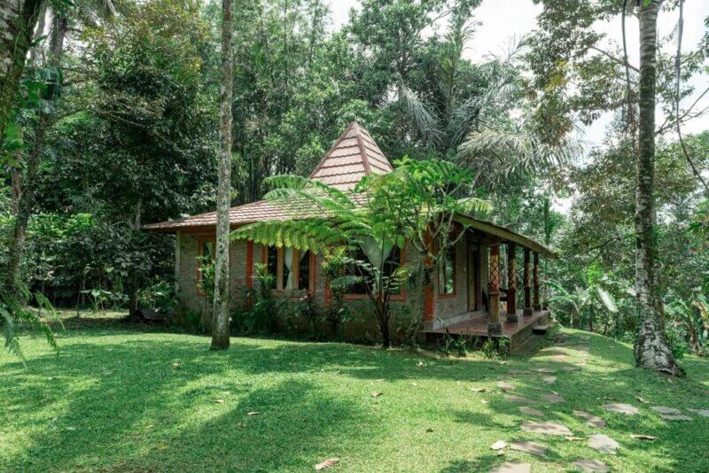 Charming small brick house with a tiled roof at Saridevi Ecolodge, nestled amid lush green grass, tropical vegetation, and trees—perfect for a tranquil Bali getaway on a sunny day.