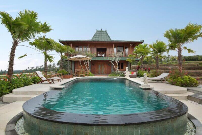 A two-story wooden villa with a green-tiled roof, surrounded by palm trees, features an infinity pool and lounge chairs in front, set in a landscaped garden.