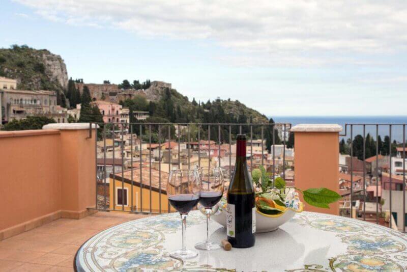 A round table with a wine bottle, two glasses of red wine, and a fruit bowl sits on the terrace of Vilagos Apartments, overlooking the stunning coastal town of Taormina and the sparkling sea.