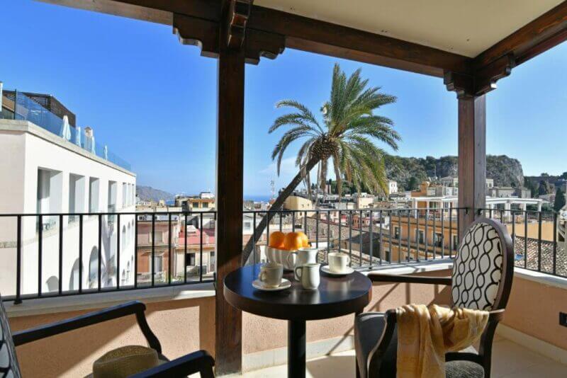 Covered balcony at Hotel Le Chevalier with a table set for coffee, two chairs, and a palm tree, overlooking Taormina’s cityscape with buildings and distant hills under a clear blue sky.