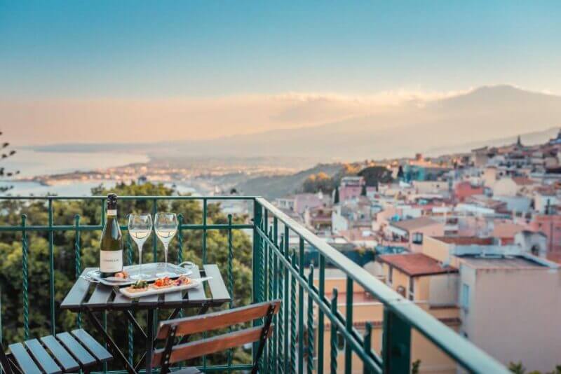 A small table with wine, glasses, and food on a balcony in Taormina overlooks the town, coastline, and Teatro Greco under a clear sky at sunset.