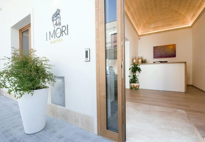 Hotel entrance with an open glass door, white exterior wall displaying I Mori Hotel sign, potted plants, and a reception desk visible inside—your welcoming stay in San Vito begins at I Mori Hotel.