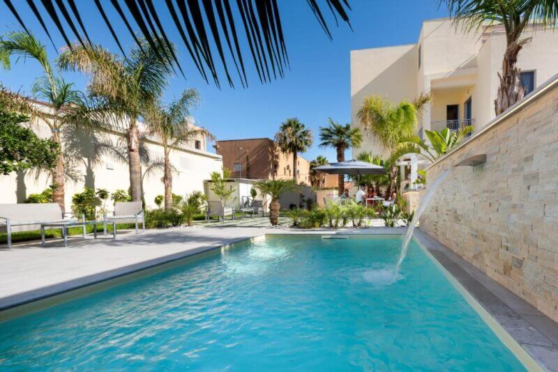 Outdoor swimming pool with a water feature, surrounded by palm trees and sun loungers at Fior di Lino, San Vito, set against modern buildings under a clear blue sky.