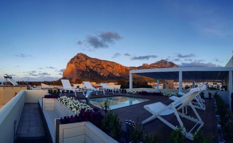 Rooftop pool area at Artemide Hotel in San Vito, featuring white lounge chairs, vibrant flowers, and a pergola, all overlooking a mountain at sunset with a partly cloudy sky.
